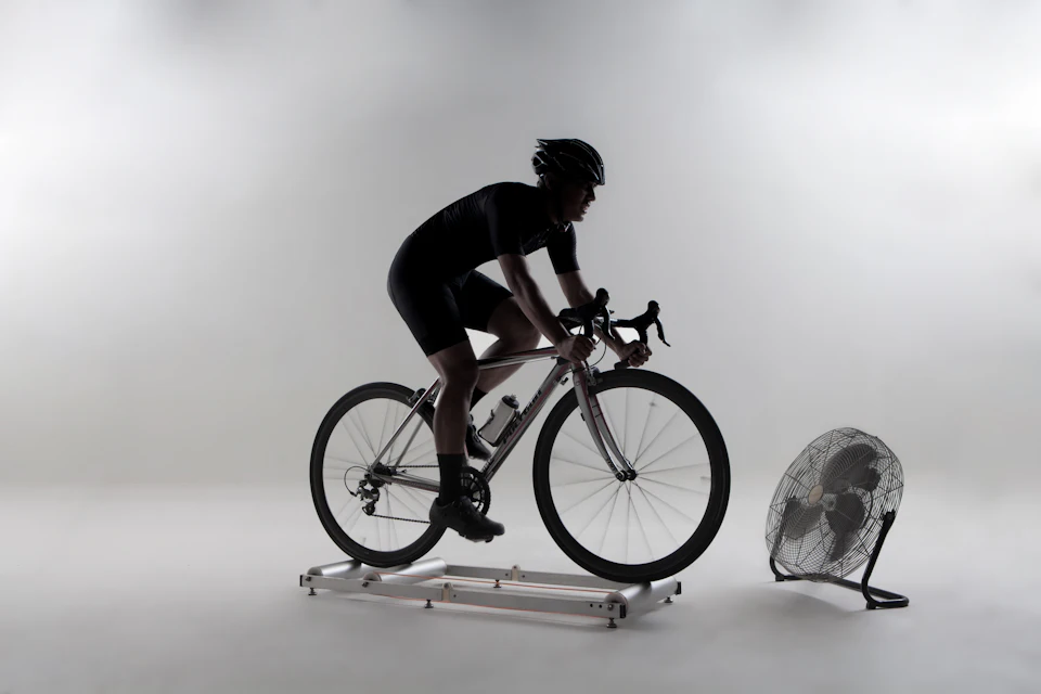 Silhouette of a cyclist training indoors on a bike fixed to a trainer, with a fan beside it