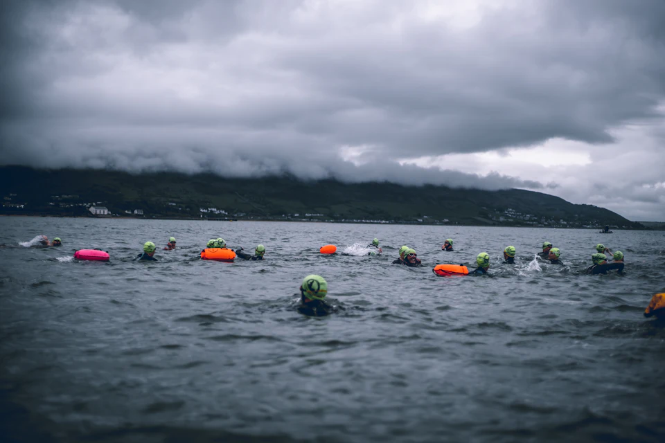 Open water swimmers in bright caps and tow floats on a lake under a stormy sky